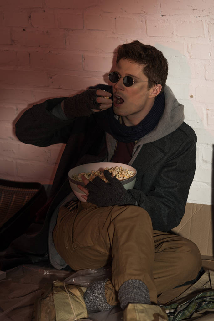πεινασμένος άστεγοι άνθρωπος χωροθέτηση με τοίχο από τούβλα και τρώγοντας ποπ κορν - Φωτογραφία, εικόνα