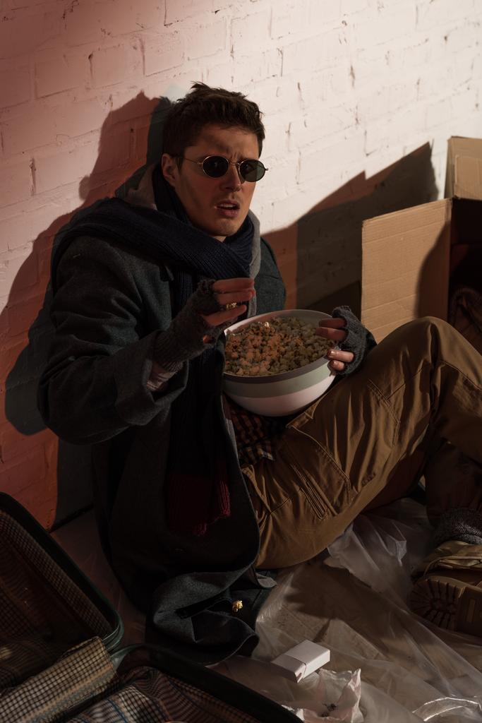hombre sin hogar sentado junto a la pared de ladrillo y sosteniendo un tazón de palomitas de maíz
  - Foto, imagen