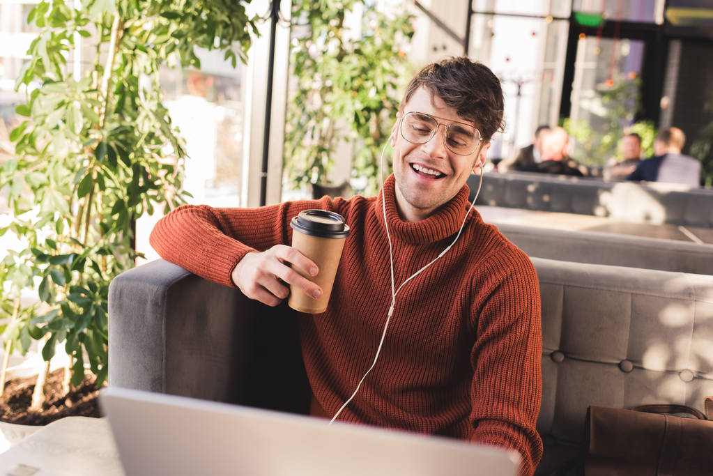 hombre sonriente escuchando música en los auriculares mientras usa el ordenador portátil y sostiene la taza desechable en la cafetería
 - Foto, imagen