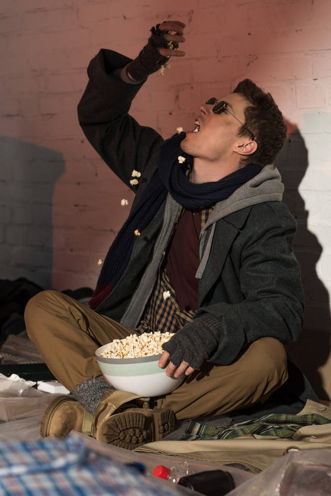 πεινασμένος άστεγοι άνθρωπος τρώει ποπ-κορν, ενώ η συνεδρίαση με τοίχο από τούβλα - Φωτογραφία, εικόνα
