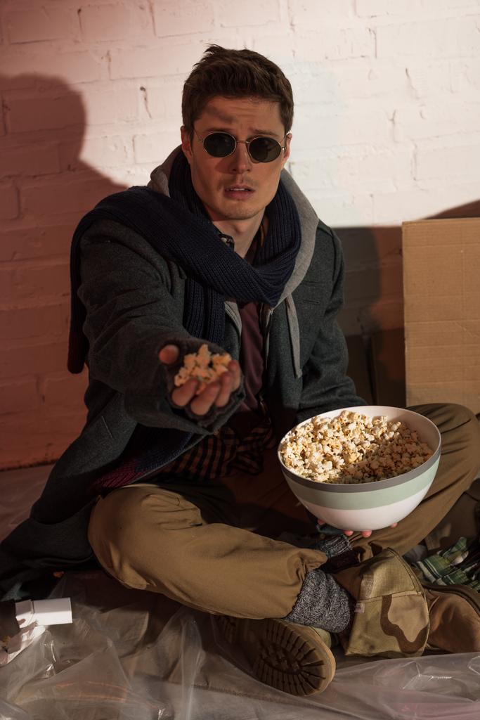 evsiz adam beyaz tuğla duvar dibinde otururken yükseltilmiş elinde patlamış mısır holding - Fotoğraf, Görsel