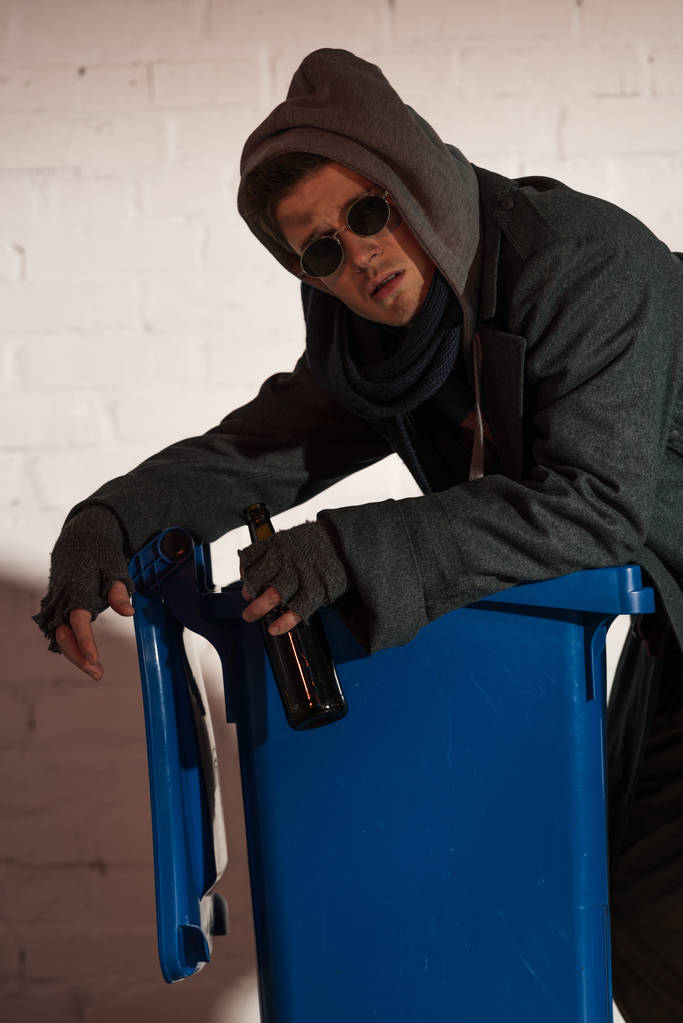 hombre sin hogar con botella de alcohol apoyada en contenedor de basura
  - Foto, Imagen