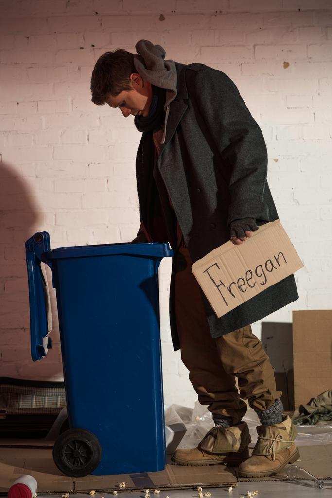 hombre sin hogar escudriñando en contenedor de basura mientras sostiene la tarjeta de cartón con letras escritas a mano "freegan"
 - Foto, Imagen