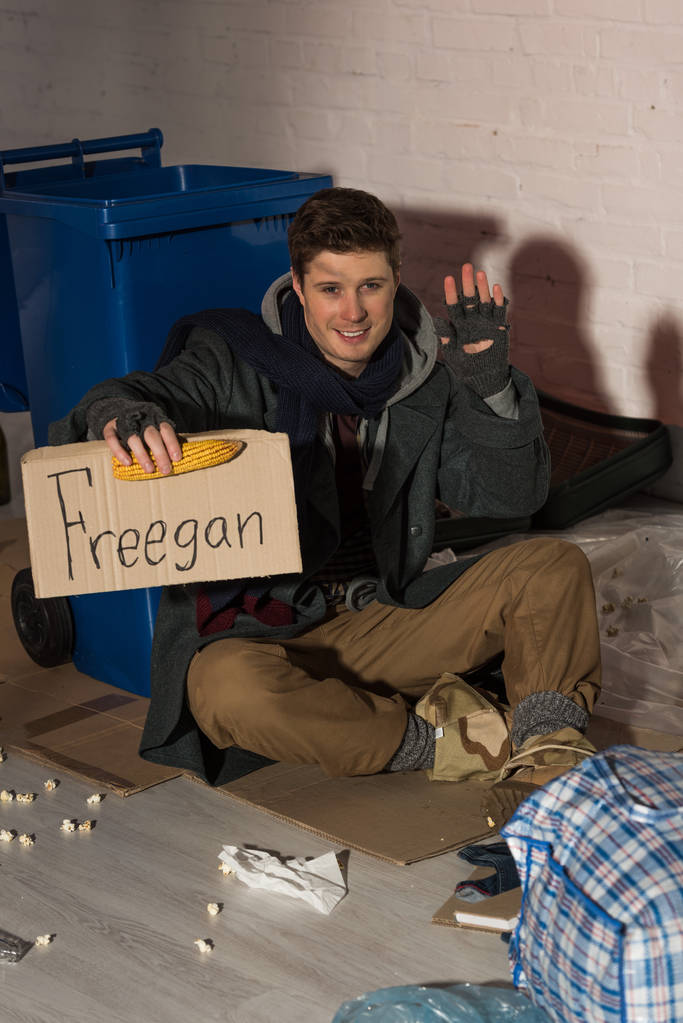 Lächelnder Obdachloser winkt mit der Hand, während er eine Pappkarte mit der Aufschrift "Freegan" und Maiskolben in der Hand hält - Foto, Bild