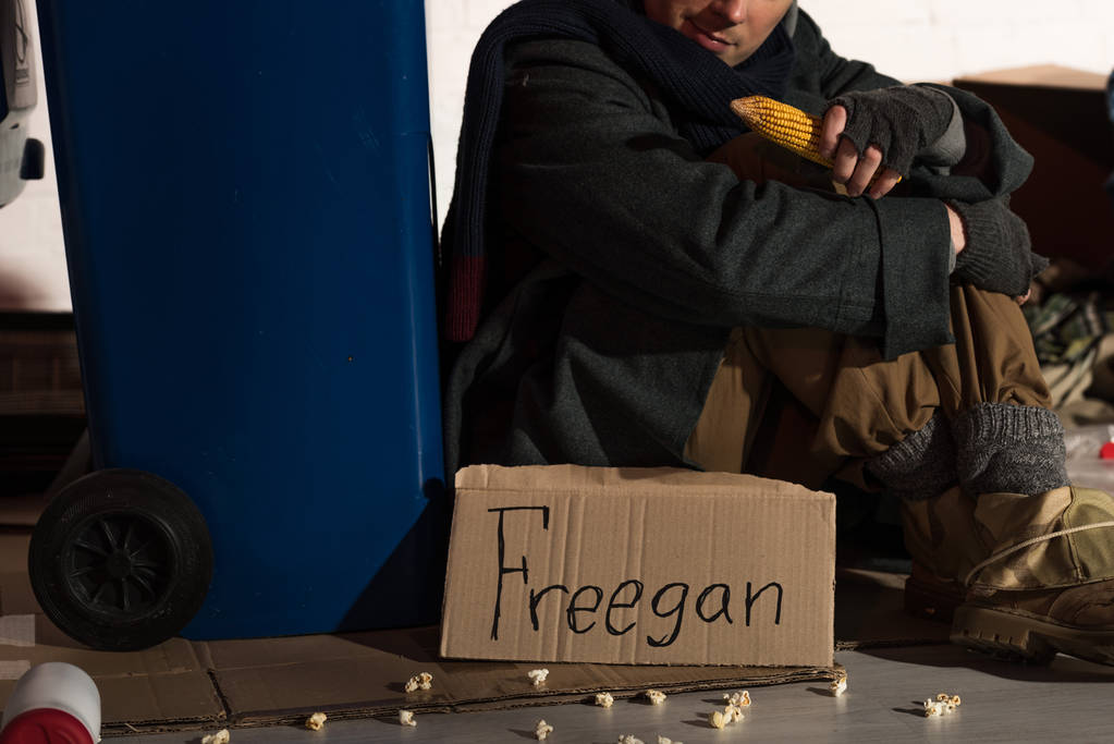 oříznutý pohled bezdomovec sedí poblíž odpadkových nádob a držel Kukuřičný klas - Fotografie, Obrázek