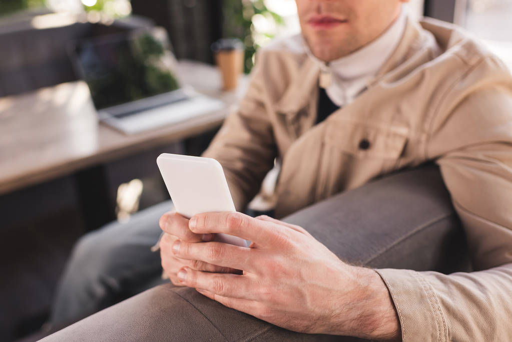 обрезанный вид на модного человека, сидящего рядом с ноутбуком и бумажной чашкой и использующего смартфон
 - Фото, изображение