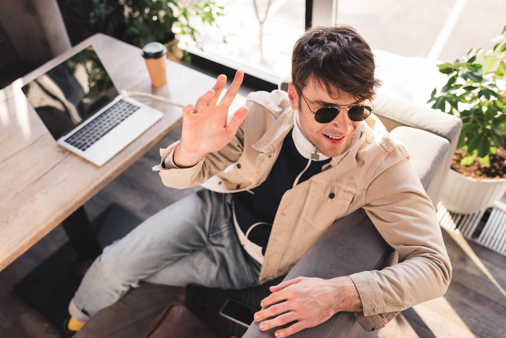 valikoiva painopiste trendikäs mies aurinkolasit istuu lähellä kannettavaa tietokonetta ja paperimukia, kun heiluttaa käsi kahvilassa
 - Valokuva, kuva