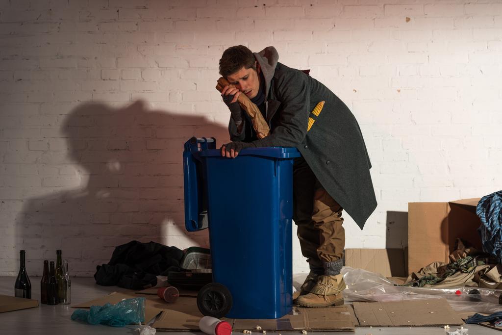 Бездомный держит хлебный багет у лица, опираясь на мусорный контейнер.
 - Фото, изображение