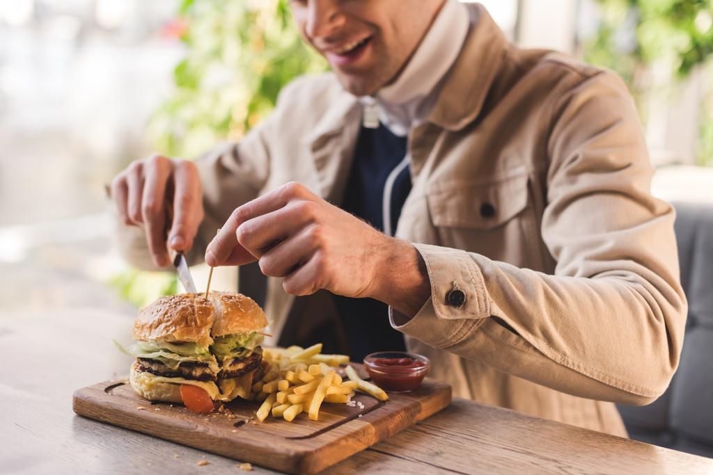 cropped view of cheerful young man cutting tasty burger on cutting board in cafe - Photo, Image