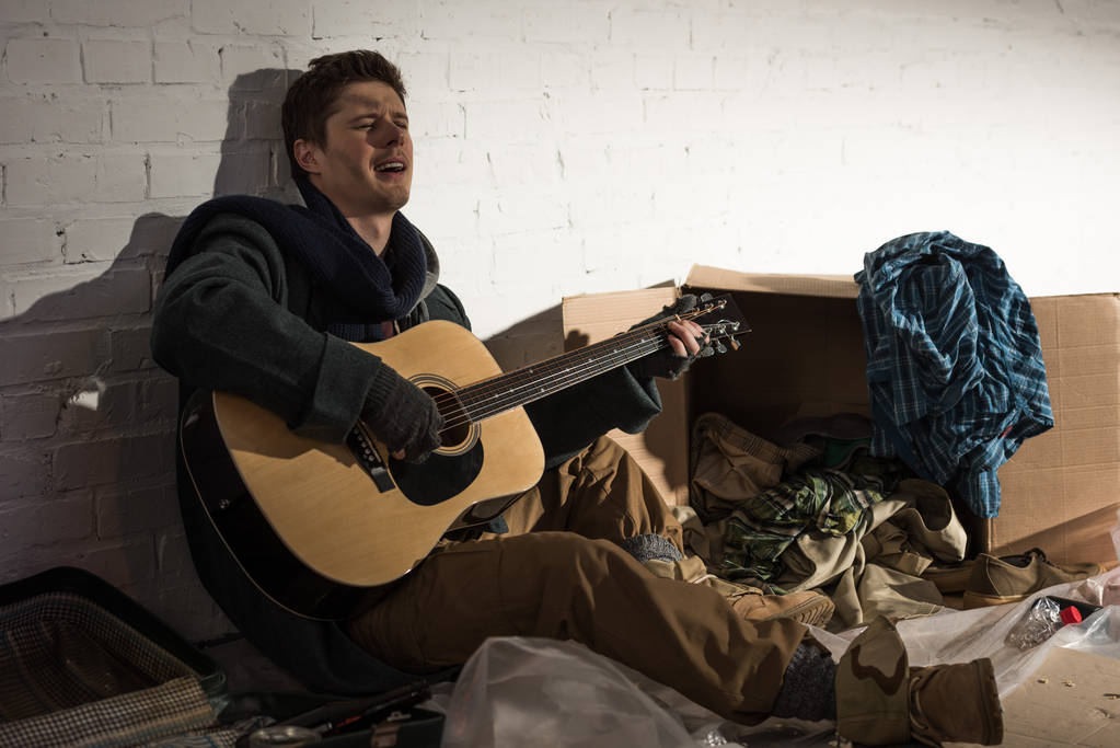 бездомний чоловік грає на гітарі та співає, сидячи без сміття
 - Фото, зображення