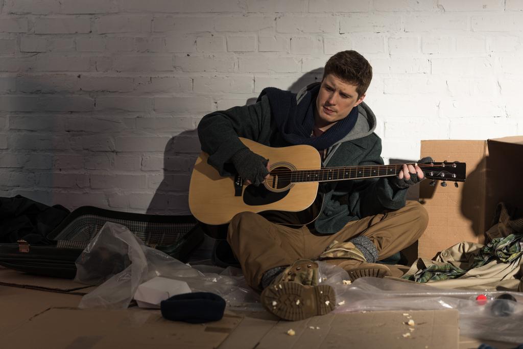 dakloze man zittend op een vuilnisbelt en gitaar spelen  - Foto, afbeelding