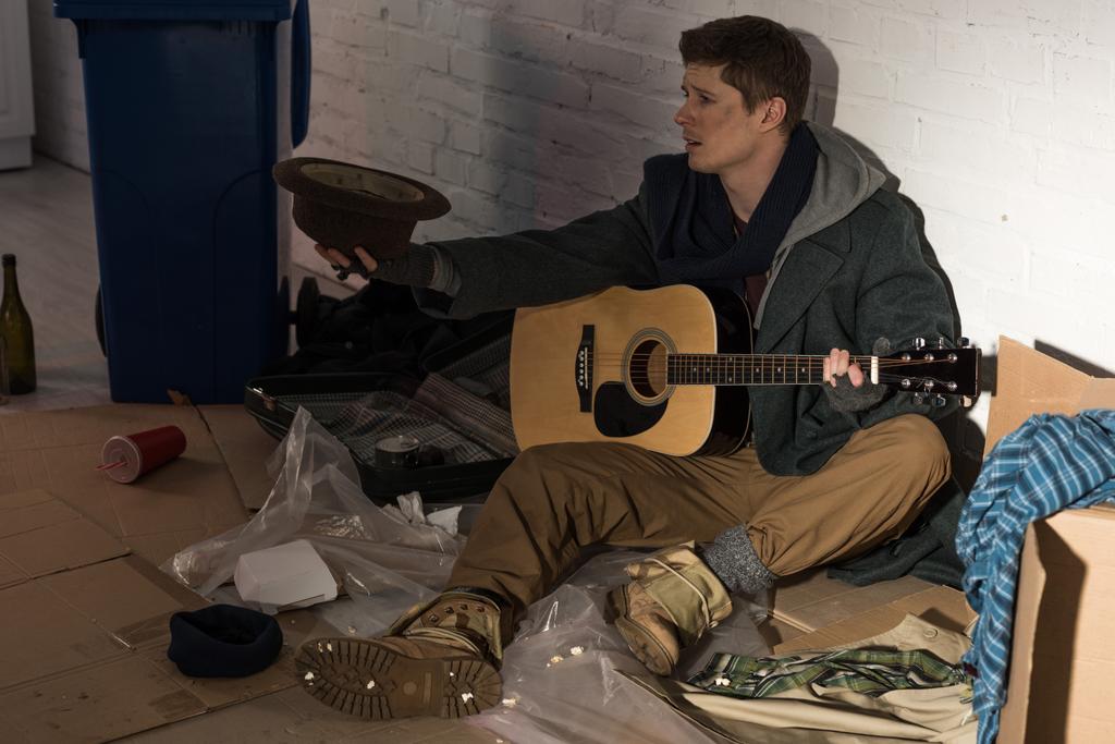 koditon mies kitara istuu kaatopaikalla ja pitelee hattua venytetyssä kädessä
 - Valokuva, kuva