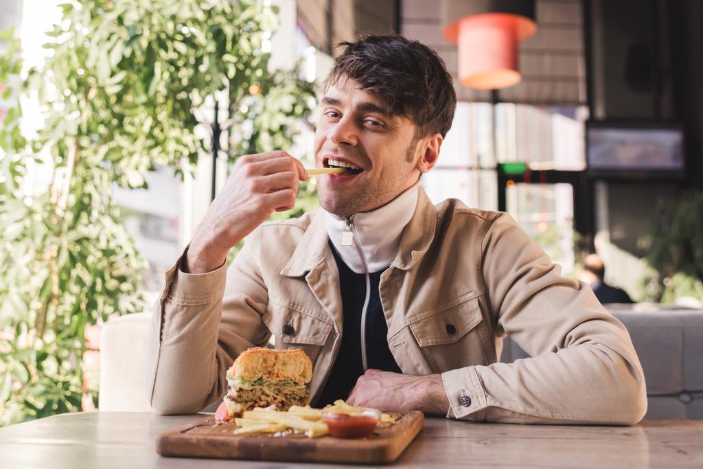 cheerful young man eating french fry near tasty burger on cutting board in cafe - Photo, Image