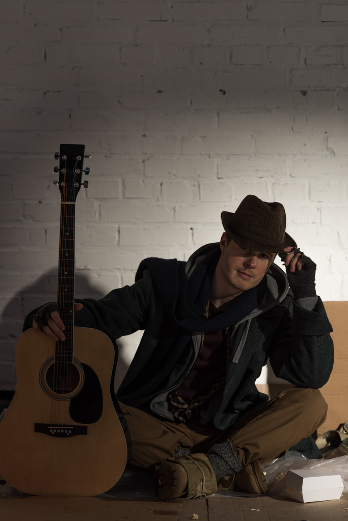 Бездомный в шляпе сидит на картонке у белой кирпичной стены и держит гитару
 - Фото, изображение