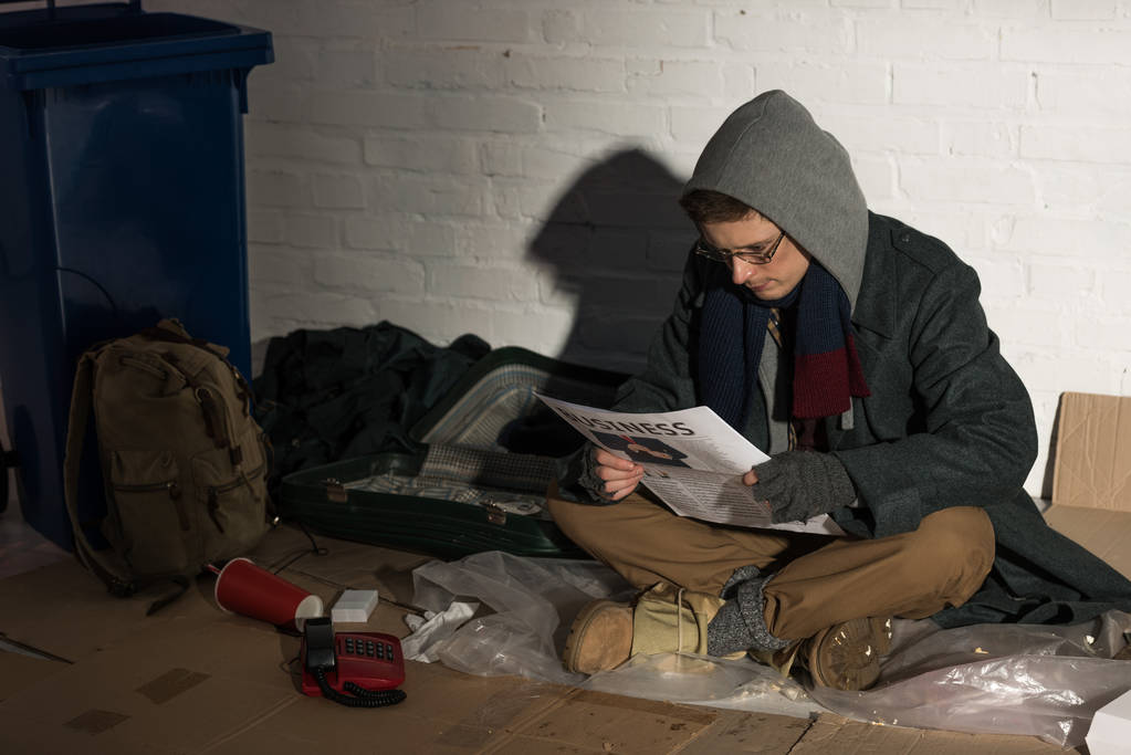 homeless man in hood and glasses reading business newspaper while sitting by brick wall - Photo, Image