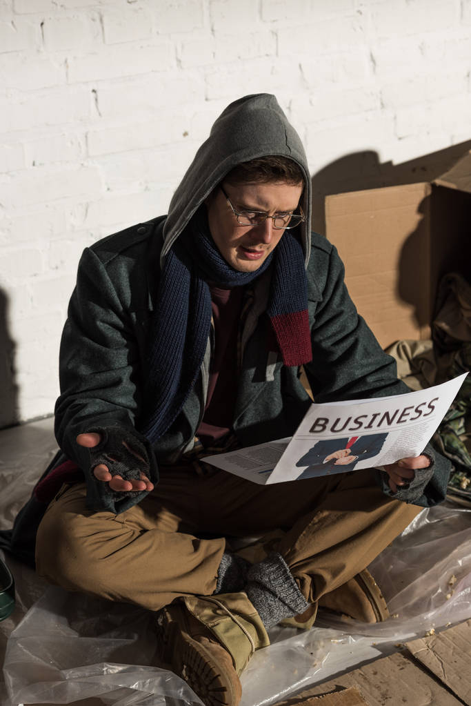 sorprendido hombre sin hogar leyendo periódico de negocios mientras estaba sentado en el basurero
 - Foto, Imagen