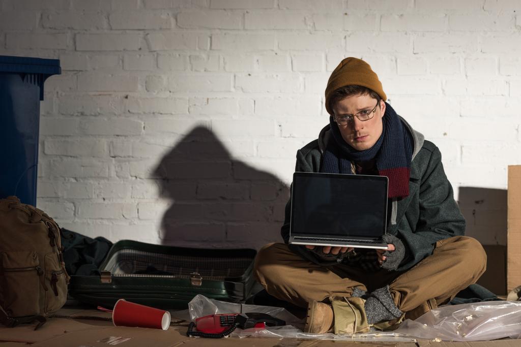 ernstige dakloze man met behulp van laptop zittend door bakstenen muur op vuilnisbelt - Foto, afbeelding
