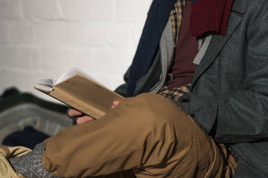 cropped view of homeless man reading book while sitting by white brick wall - Photo, Image