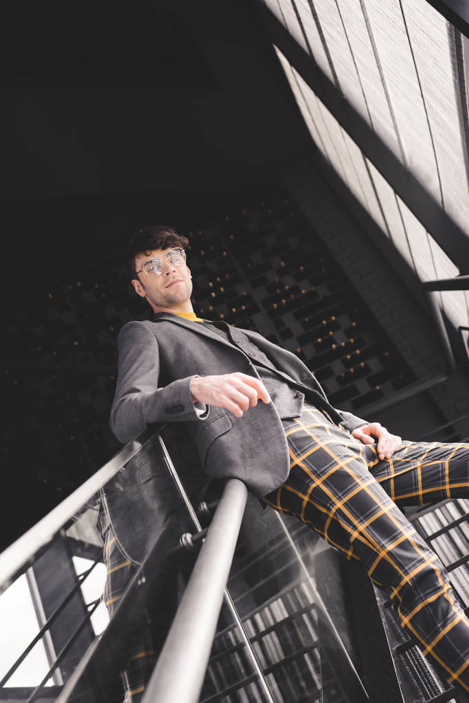 young stylish man in glasses posing on stairs and looking at camera - Photo, Image