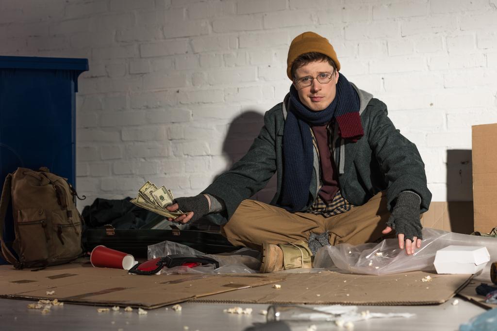 Бездомный держит деньги, сидя на мусорной свалке
 - Фото, изображение