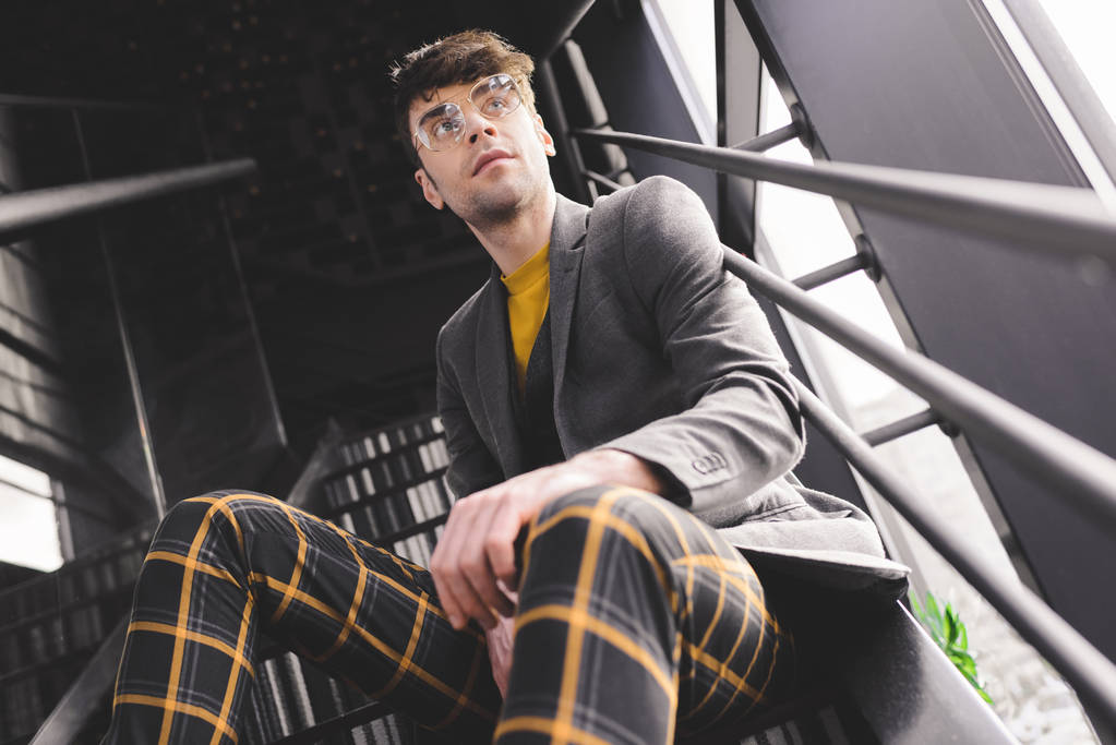 мечтательный стильный мужчина в очках сидит на лестнице возле окон
 - Фото, изображение