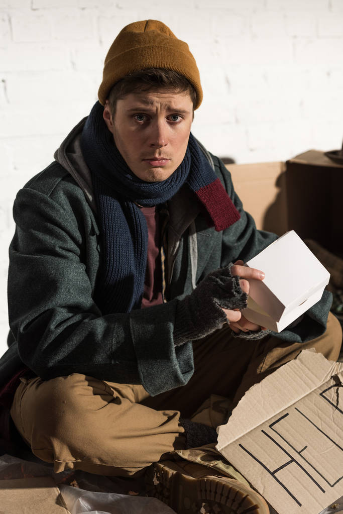 печальный бездомный сидит с картонной карточкой с надписью "Помогите" и держит белую бумажную коробку
 - Фото, изображение