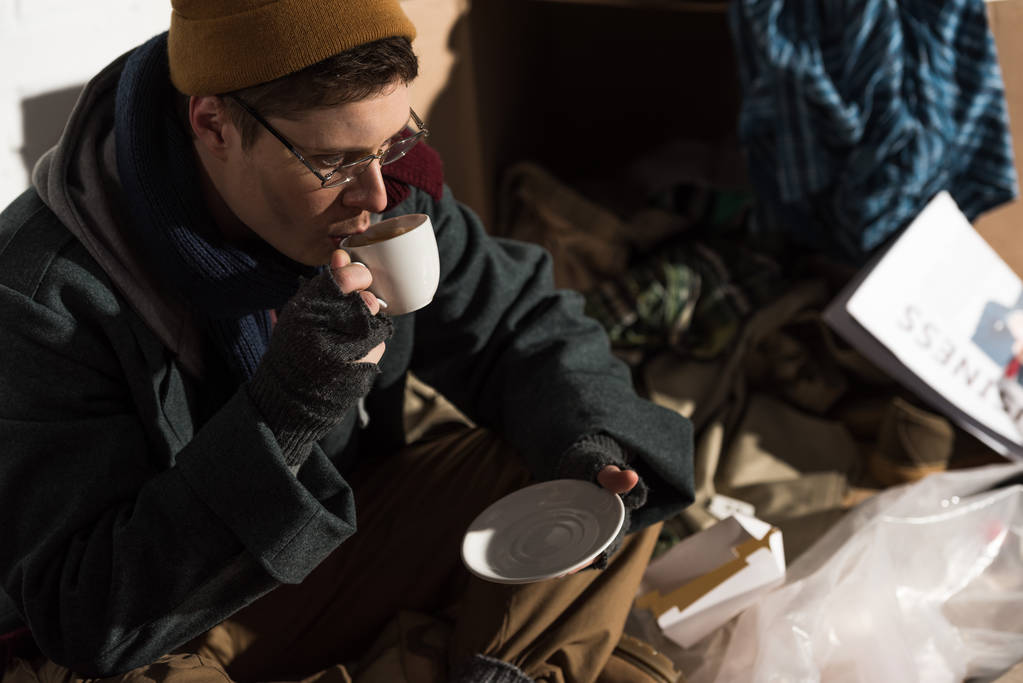 Бездомный в очках и перчатках без пальцев пьет кофе
 - Фото, изображение