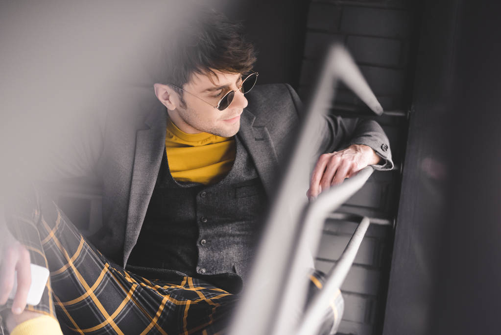 вибірковий фокус красивого чоловіка в сонцезахисних окулярах, що сидять на сходах зі смартфоном в руці
  - Фото, зображення