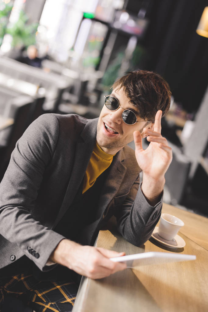 happy young man in sunglasses gesturing near bar counter while holding digital tablet near cup with coffee - Photo, Image