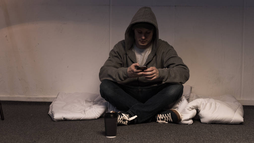ο άνθρωπος σε hoodie χρησιμοποιώντας smartphone, ενώ κάθεται στο στρώμα που τακτοποιείται στον όροφο - Φωτογραφία, εικόνα