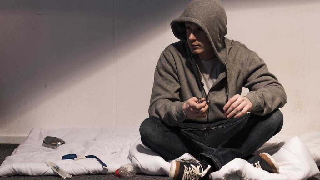 наркоман в толстовке держит трубку для курения, сидя на матрасе в темной комнате
 - Фото, изображение
