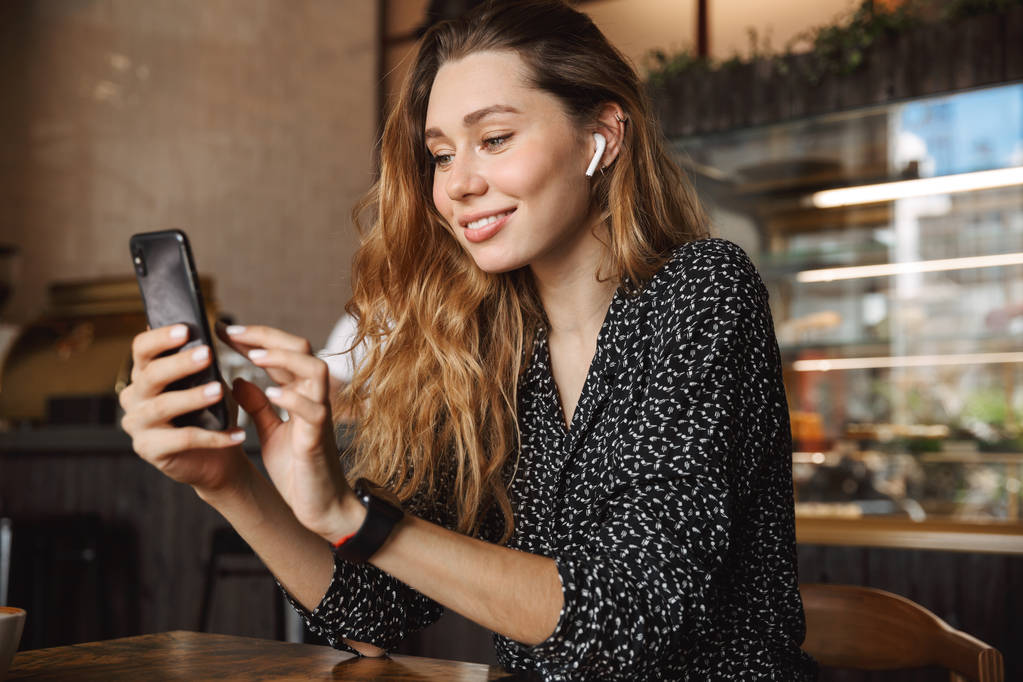 Φωτογραφία: χαρούμενα ευτυχής όμορφη νεαρή όμορφη γυναίκα κάθεται στο καφενείο indoors χρησιμοποιώντας κινητό τηλέφωνο, ακούτε μουσική με ακουστικά. - Φωτογραφία, εικόνα