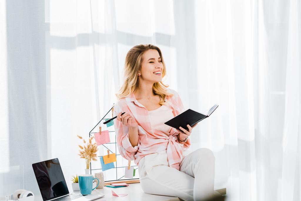 Laughing young woman with notebook sitting on table at home office - Photo, Image