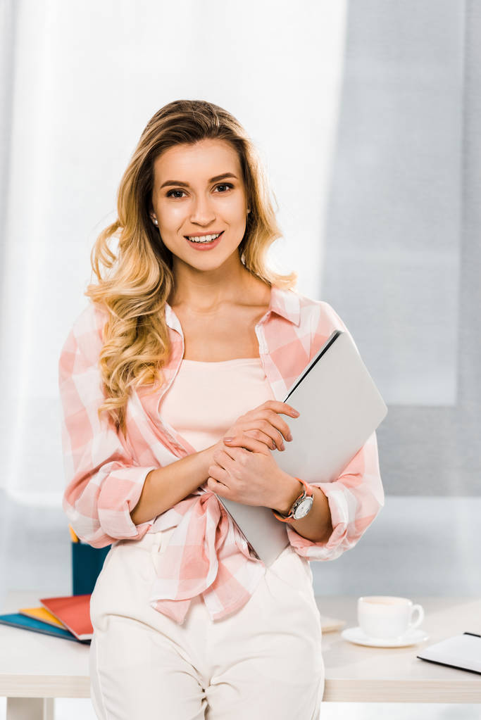 hinreißend lockige Frau im karierten Hemd hält Laptop in der Hand und blickt lächelnd in die Kamera - Foto, Bild