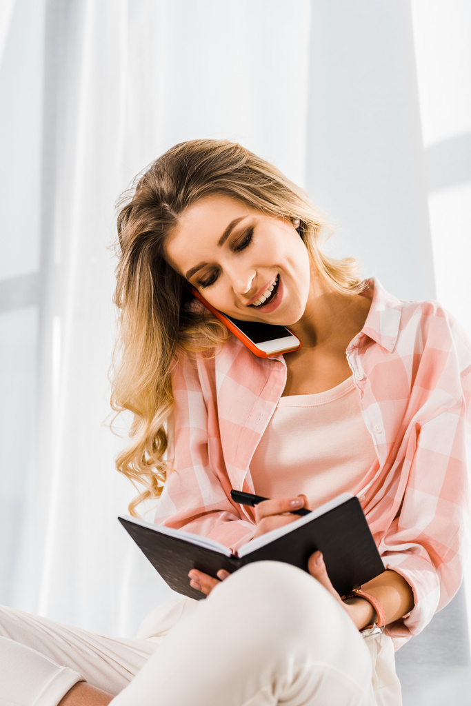 Lachende vrouw in geruite overhemd op smartphone praten en schrijven in notitieblok - Foto, afbeelding