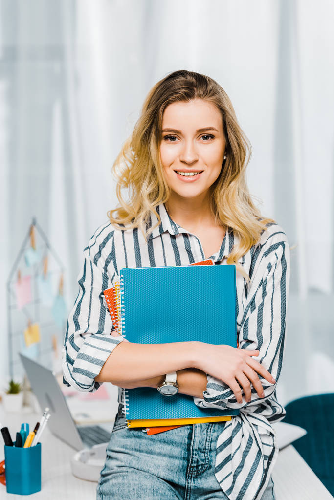 Glad young woman in striped shirt holding notebooks and looking at camera with smile - Photo, Image