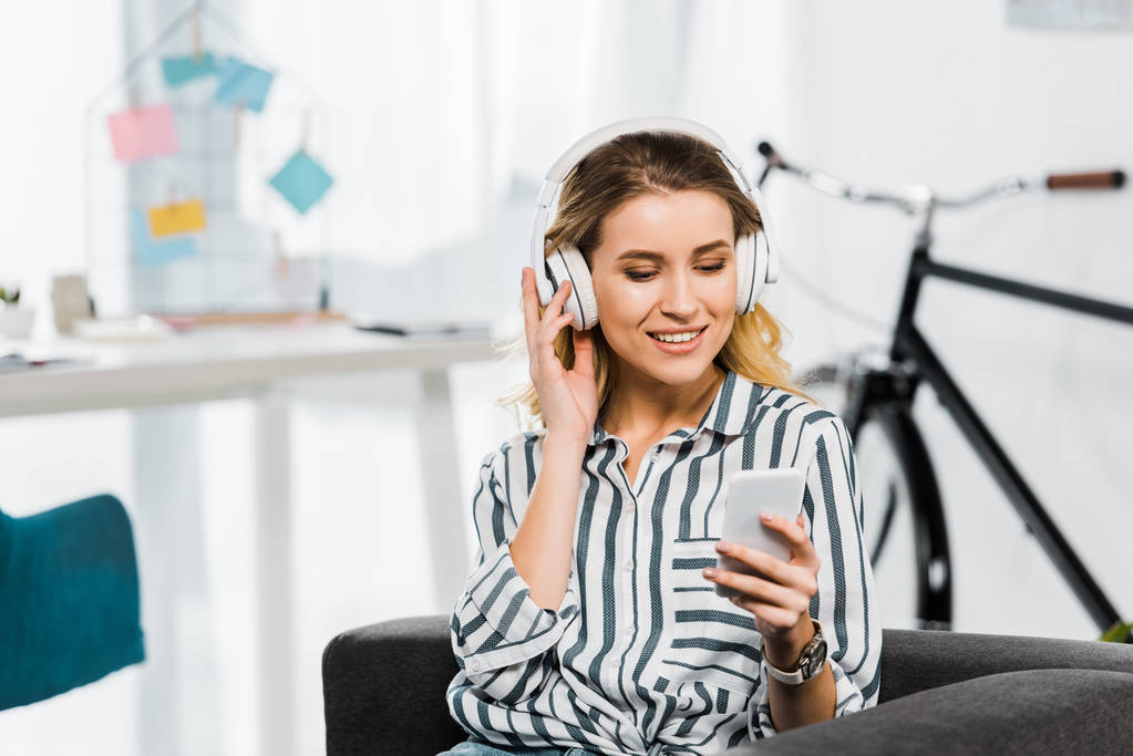 Mujer joven feliz con camisa a rayas escuchando música y sosteniendo el teléfono inteligente
 - Foto, imagen
