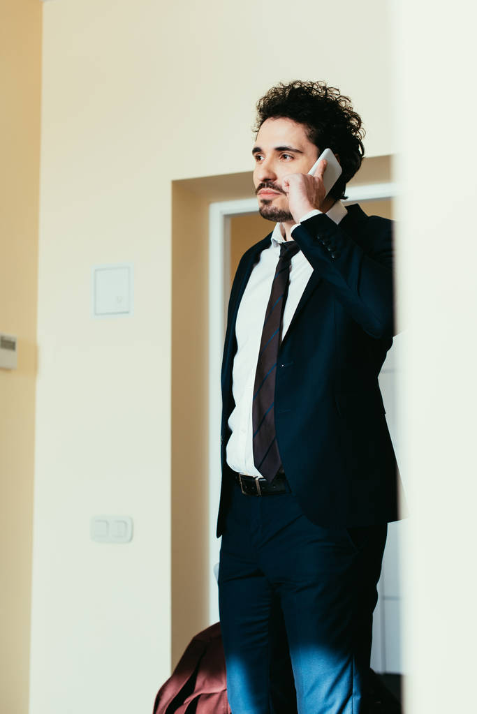 ホテルの部屋でビジネス旅行中にスマート フォンで話しているスーツのビジネスマン - 写真・画像