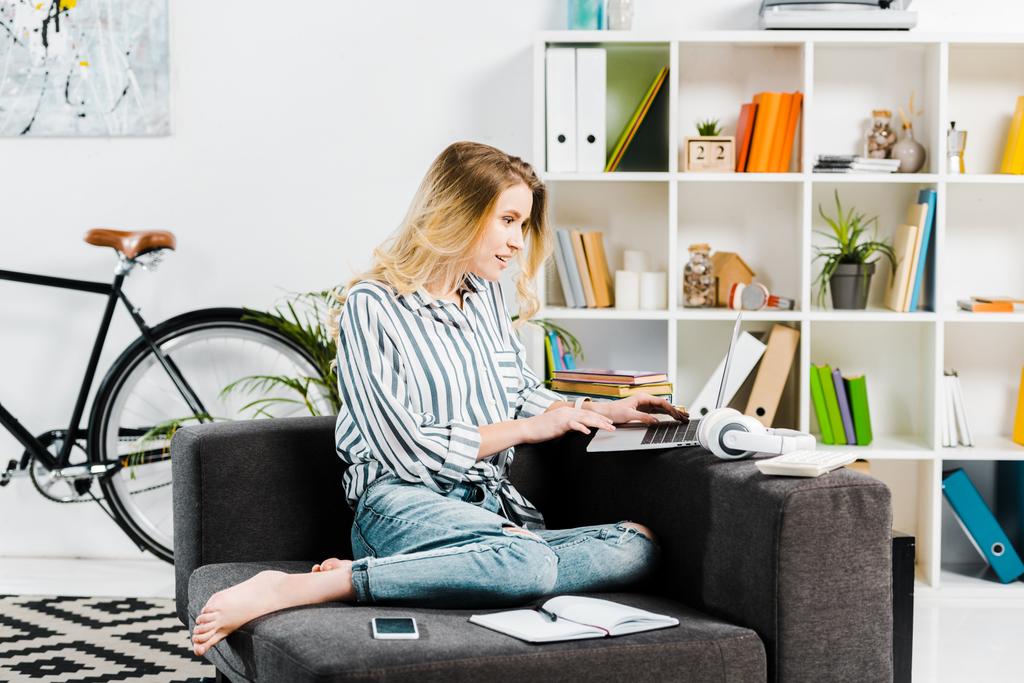 Jolie femme en chemise rayée assise sur un canapé et utilisant un ordinateur portable
 - Photo, image