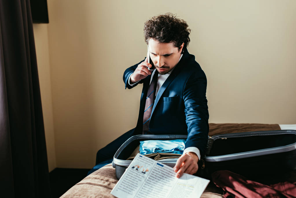 бізнесмен розмовляє на смартфоні і тримає газету, сидячи на ліжку з валізою в готельному номері
 - Фото, зображення