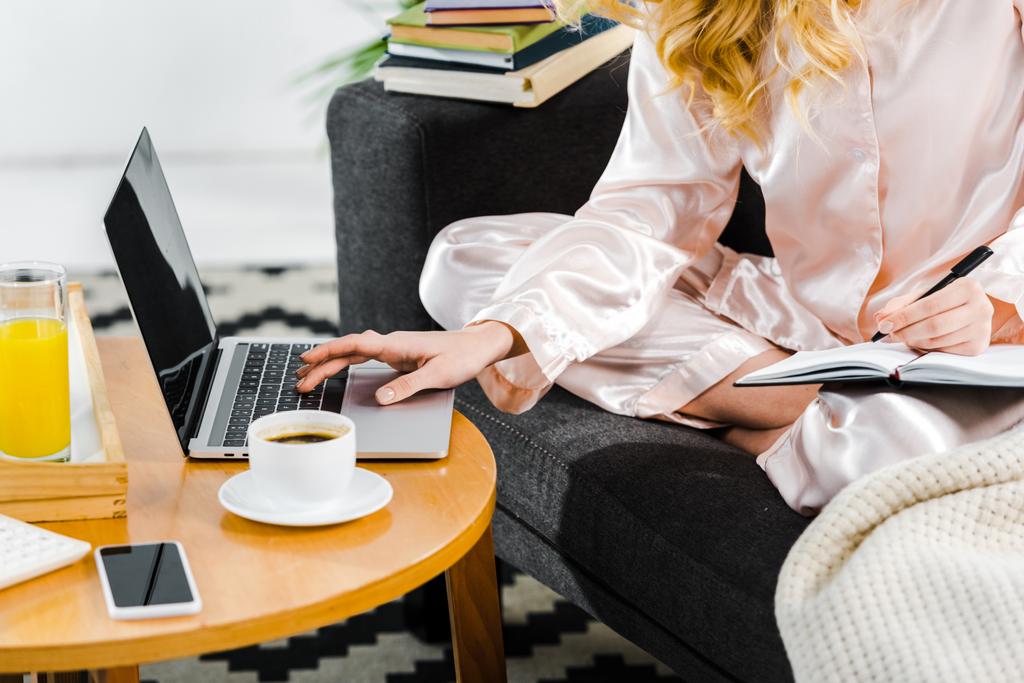 Vista parziale della donna in pigiama seduta sul divano e che utilizza laptop con schermo bianco
 - Foto, immagini