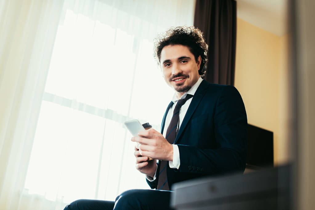 hymyilevä liikemies käyttää älypuhelinta työmatkalla hotellihuoneessa
 - Valokuva, kuva