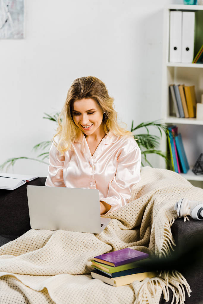 Pijamalı kız kanepede oturan ve dizüstü bilgisayar kullanarak ekose ile gülümseyen - Fotoğraf, Görsel