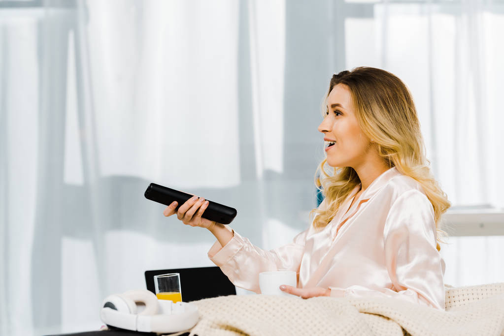 Smiling woman in pyjamas holding coffee cup and tv remote control - Photo, Image