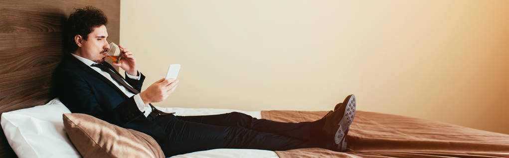 homme d'affaires en costume en utilisant smartphone et boire du whisky sur le lit dans la chambre d'hôtel
 - Photo, image