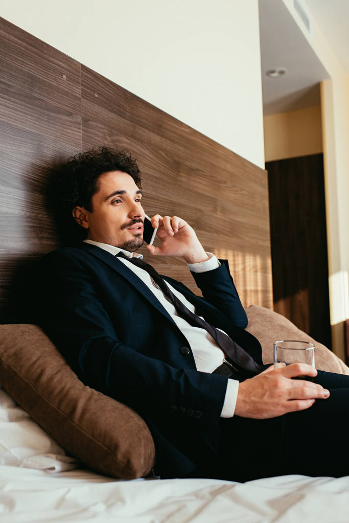 hombre de negocios hablando en el teléfono inteligente y sosteniendo vaso de whisky en la cama en la habitación de hotel
 - Foto, Imagen