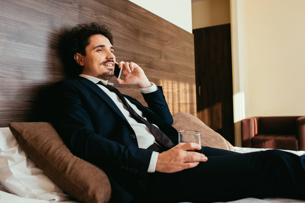 улыбающийся бизнесмен разговаривает по смартфону и держит стакан виски на кровати в гостиничном номере
 - Фото, изображение