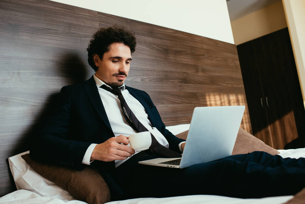 hombre de negocios en traje sosteniendo la taza de café y trabajando en el ordenador portátil en la habitación de hotel
 - Foto, imagen