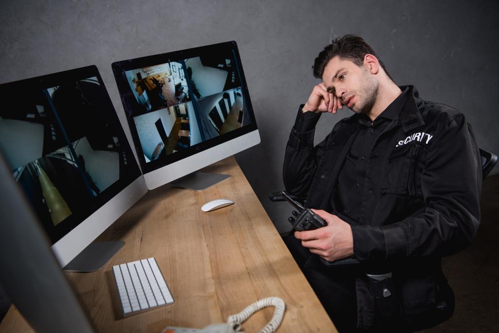 скучающий охранник в форме держит рацию и смотрит на монитор компьютера
  - Фото, изображение