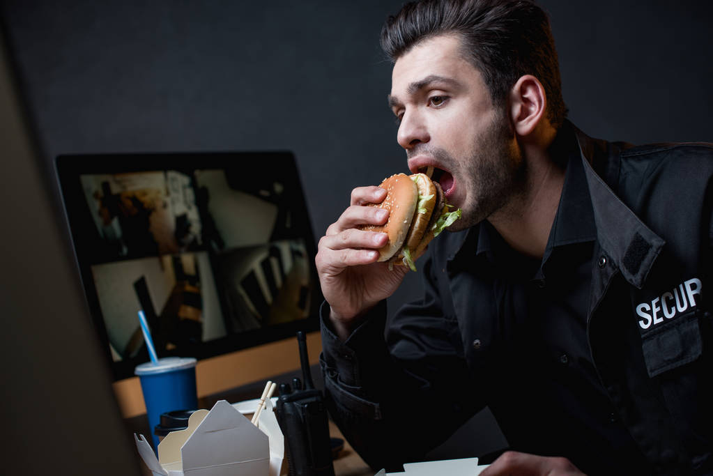Φύλαξη σε ομοιόμορφο τρώει burger και κοιτάζοντας οθόνη υπολογιστή  - Φωτογραφία, εικόνα